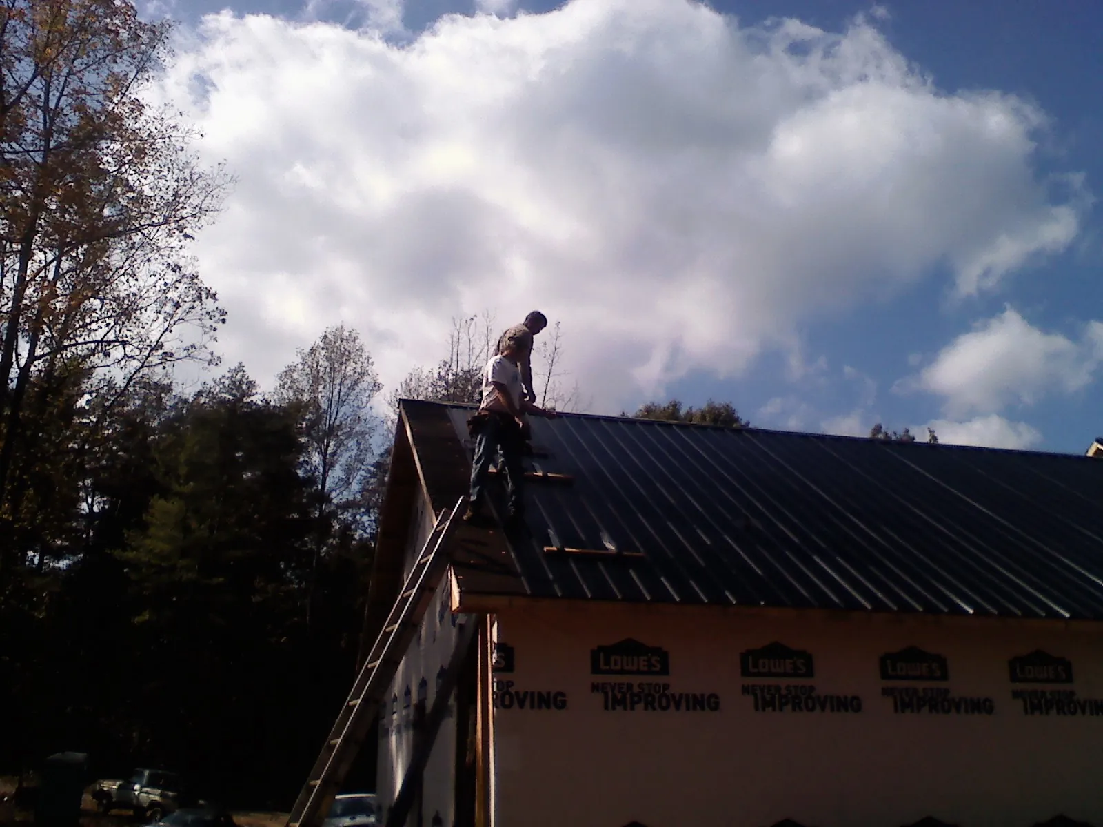 Roofer fastening dark metal panels high against a cloudy sky, from a Roof Inspection job in Raynham